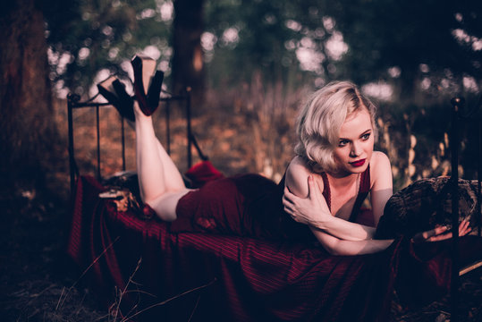 Beautiful and elegant blonde woman with red lips and hair waves wearing wine red nightie posing on the bed outdoors autumn, retro vintage style and fashion.