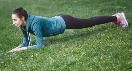 Fototapeta na wymiar Portrait of young and sporty woman in sportswear doing yoga or Stretching exercises outside at the park on green field on cloudy day, Dnipro, Ukraine. She is sitting on the green meadow between trees