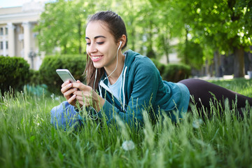 Portrait of young and sporty woman in sportswear sitting with the smartphone and headphones on the grass outside at the park on green field on cloudy day, Dnipro, Ukraine. She is Lies on the green
