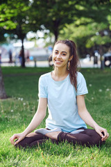 Portrait of young and sporty woman in sportswear doing yoga or Stretching exercises outside at the park on green field on cloudy day, Dnipro, Ukraine. She is sitting on the green meadow between trees