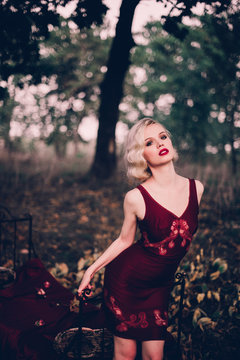 Beautiful and elegant blonde woman with red lips and hair waves wearing wine red nightie posing on the bed outdoors autumn, retro vintage style and fashion.