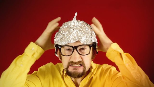 An angry ugly man wearing a tin foil hat (made from aluminium, worn in the belief or hope that it shields the brain from threats such as electromagnetic fields, mind control or reading).
