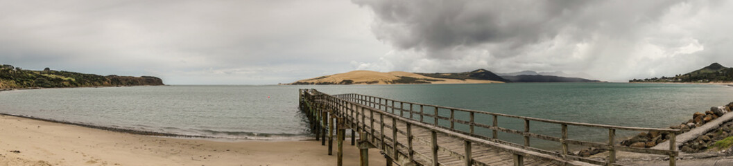 Bay of Islands, New Zealand - March 7, 2017: Panorama shot of exit to Tasman Sea from Hokianga Harbour shows large dune, pier, mountains, all under heavy cloudscape because of approaching cyclone.