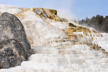 Mammoth Hot Springs with travertine terraces in foreground, the black Devil's Thumb on the left and steam rising from the water at the top. Photographed in Yellowstone National Park in natural light.