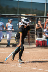 Teenage batter in black softball uniform dropping her bat after contact.  