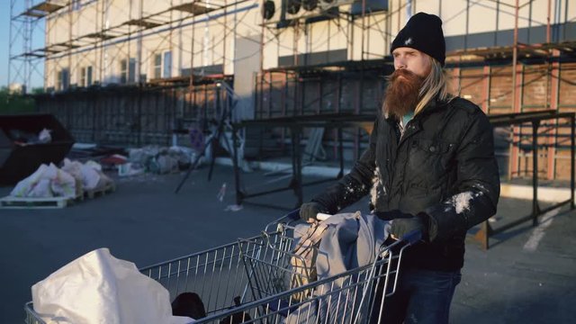 Dolly shot of bearded homeless man walking down the street with shooping card at cold autumn day