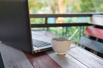 white ceramic coffee cup have spoon inside putting on beside laptop computor all both putting on wooden table. nature are background. this image for business and technology concept