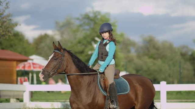 SLOW MOTION CLOSE UP: Young girl horseback riding beautiful chestnut mare in outdoor riding arena during the sunny summer vacations. Horse with a child rider trotting in sandy manege inside the corral