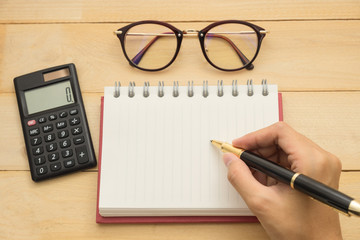 top view. hand using ballpoint pen writing on empty notebook paper have calculator put on beside and have glasses put on top. wooden are background. this image for business and education concept
