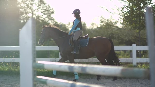 SLOW MOTION CLOSE UP: Young girl horseback riding beautiful chestnut mare in outdoor riding arena during the sunny summer vacations. Horse with a child rider trotting in sandy manege inside the corral