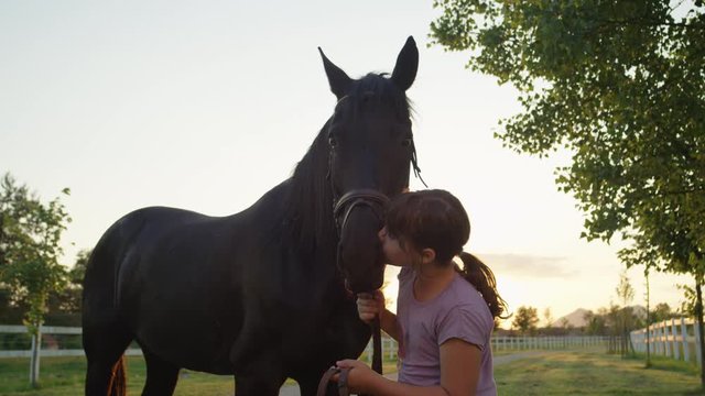 SLOW MOTION, CLOSE UP: Portrait of a cheerful little girl kissing and petting her dark bay mare at magical golden light sunset. Happy kid hugging and cuddling a colt at the countryside horse ranch