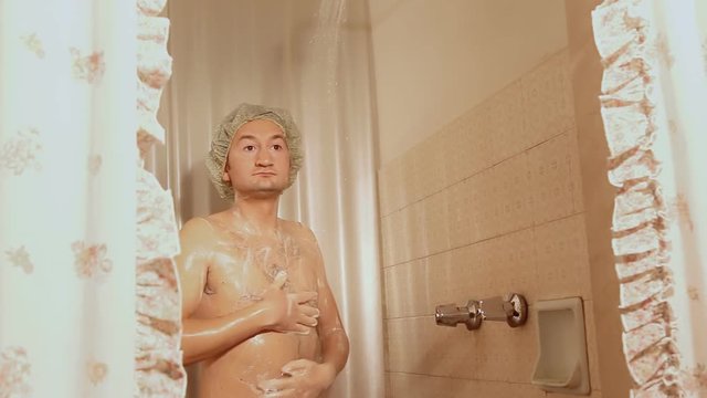 A funny slim ugly man singing in the shower, soaping himself while wearing a cap. Someone else is inside the tub, peeping at him. Gotcha! Close-up shot.
