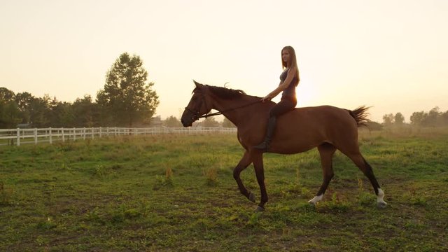 SLOW MOTION, CLOSE UP: Beautiful young Caucasian girl with long blonde hair bareback riding stunning brown horse on meadow field on misty morning at golden sunrise. Relaxing evening ride at sunset