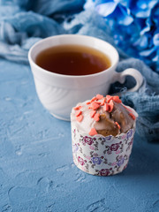 Cup of tea on blue background with frosted cup cake, flowers and textile