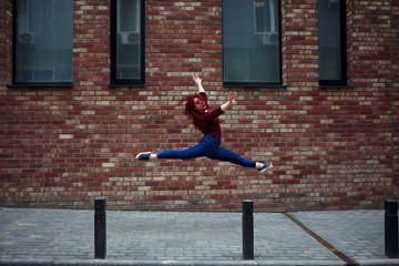 Beautiful serious young Caucasian girl dancer jumping against brick wall In jeans and a burgundy...