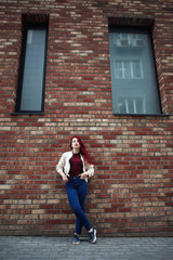 Fototapeta na wymiar Young serious caucasian woman looking at camera outdoor against brick wall on the street. wearing jeans and a burgundy shirt and white jacket with red lips full length portrait.
