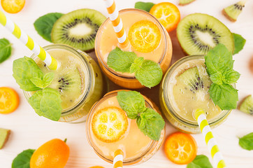 Freshly blended orange kumquat and green kiwi fruit smoothie in glass jars with straw, mint leaf, cut ripe berry, top view, close up. White wooden board background.