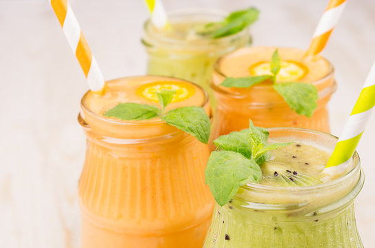Freshly blended orange kumquat and green kiwi fruit smoothie in glass jars with straw, mint leaf, cut ripe berry, top view, close up. White wooden board background.