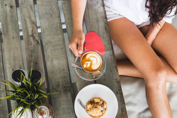Tanned woman sitting and holding cup of aroma cappuccino in hands. Tasty muffin on wooden table for...