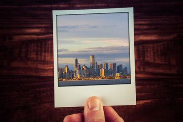 Polaroid postcard photograph of Melbourne skyline from waterfront held by male hand over wooden...
