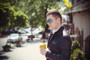Young brunette man walk in center of city and European streets in leather black jacket and blue mirror sunglasses. Student meeting with friend and wait him