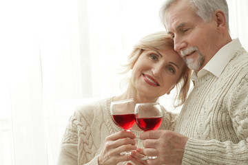 Cute elderly couple drinking wine while standing near window at home