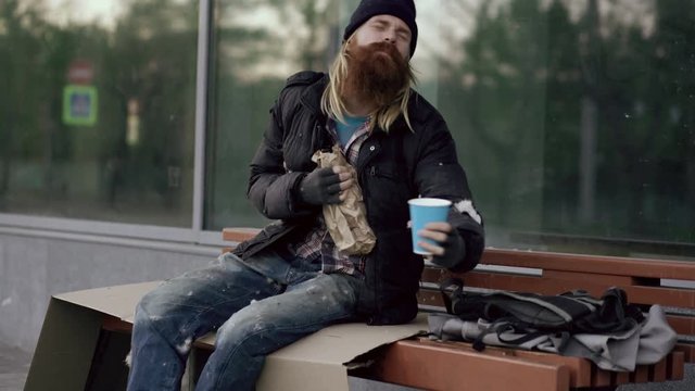 Very drunk homeless man talking to people walking near him and beg for money and show fuck you sign while sitting on bench at the sidewalk