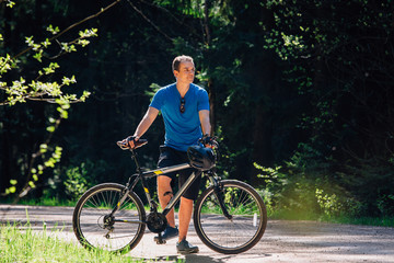 Naklejka premium Young man in blue shirt standing with bicycle on the forest road. Healthy lifestyle concept. Sports guy with bike.