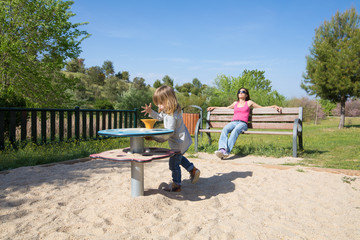 three years old blonde child playing with sand in playground next to mother sitting in bench...