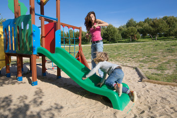 Fototapeta na wymiar mother surprised by child sliding in green plastic slide. Kid is three years old blonde. Outdoor playground, in public Park Valdebebas, Madrid Spain 