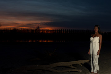 Ethereal sunset. Black model in a white dress stands on a dark beach with a sunset behind her
