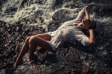 Young woman in white shirt and bikini lies on rock near waterfal