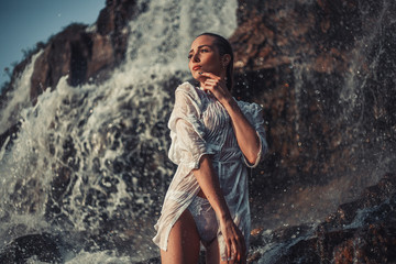 Young woman in white shirt and bikini stands near waterfall.