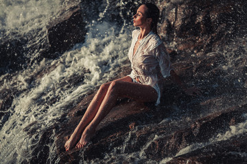 Young woman in white shirt and bikini sits on rock in water flow