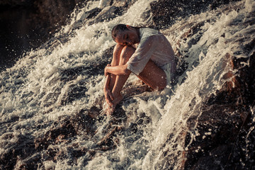 Young woman in white shirt and bikini sits on rock in water flow