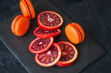 Whole and slices of blood oranges and macarons with chocolate ganache filling on a black slate...