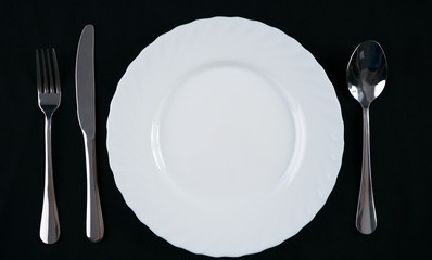 Empty white plate with silver fork, knife and spoon isolated on black background. Dinner place setting. Top view