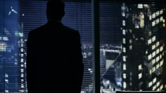 Late at Night Businessman Stands at the Window in His Office with Big City View.  Shot on RED EPIC-W 8K Helium Cinema Camera.