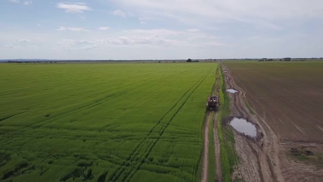 Aerial view of tractor with crop sprayer on countryside road preparing to spray insecticide and fungicide on green wheat field, drone pov