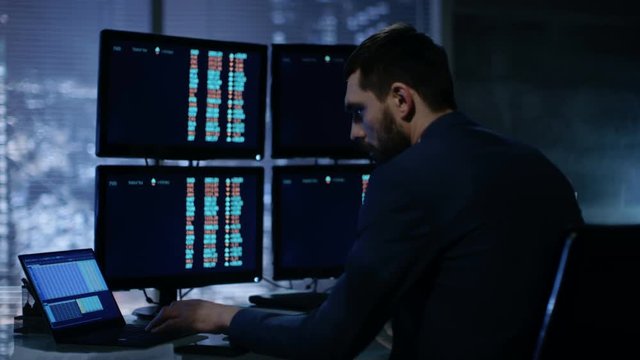 Late at Night Trader Reads Number on His Multiple Displays with Stock Information on Them. In Background Big City Window View. Shot on RED EPIC-W 8K Helium Cinema Camera.