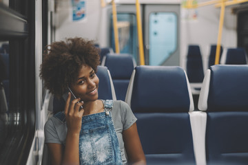 Cheerful young black lady talking with her sister via smartphone while sitting in coach of electric...