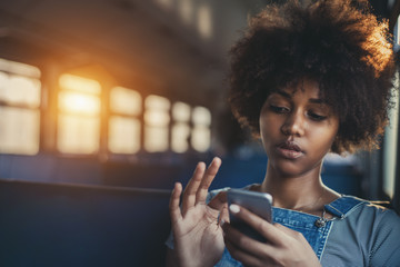 Thoughtful young biracial girl using smart phone while sitting alone in suburban train, curly african teenage female having online chat with her sister in train with copy space for your text or logo