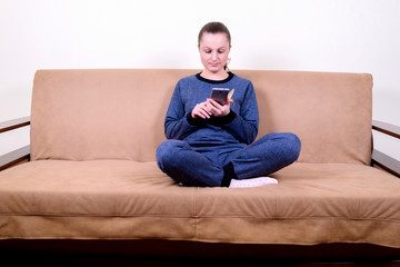 Fototapeta premium Beautiful young woman sitting on a sofa, using a smartphone and texting. Relaxing on the couch at home. Technology and communication concept