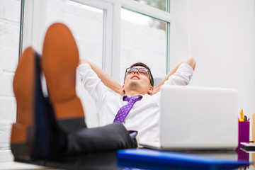 Young businessman lies on a chair and keeps legs on a table at the office