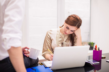 Young businesswoman writes something sitting at the table on which young businessman sits with cup of coffee at the office