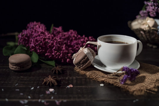 Cup of coffee on linen napkin and several macarons in braid vase with branch of lilac