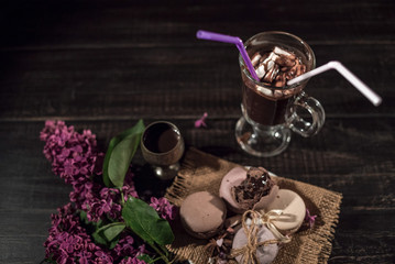 Hot chocolate (cocoa) in a glass mug with two tubes for drinking.There are some macarons on a linen napkin on the wood table with some lilac.