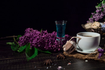 Obraz na płótnie Canvas Cup of coffee on linen napkin and several macarons in braid vase with branch of lilac and pile of absinthe