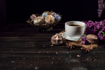 Cup of coffee on linen napkin and several macarons in braid vase with branch of lilac.
