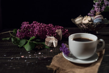 Cup of coffee on linen napkin and several macarons in braid vase with branch of lilac.
